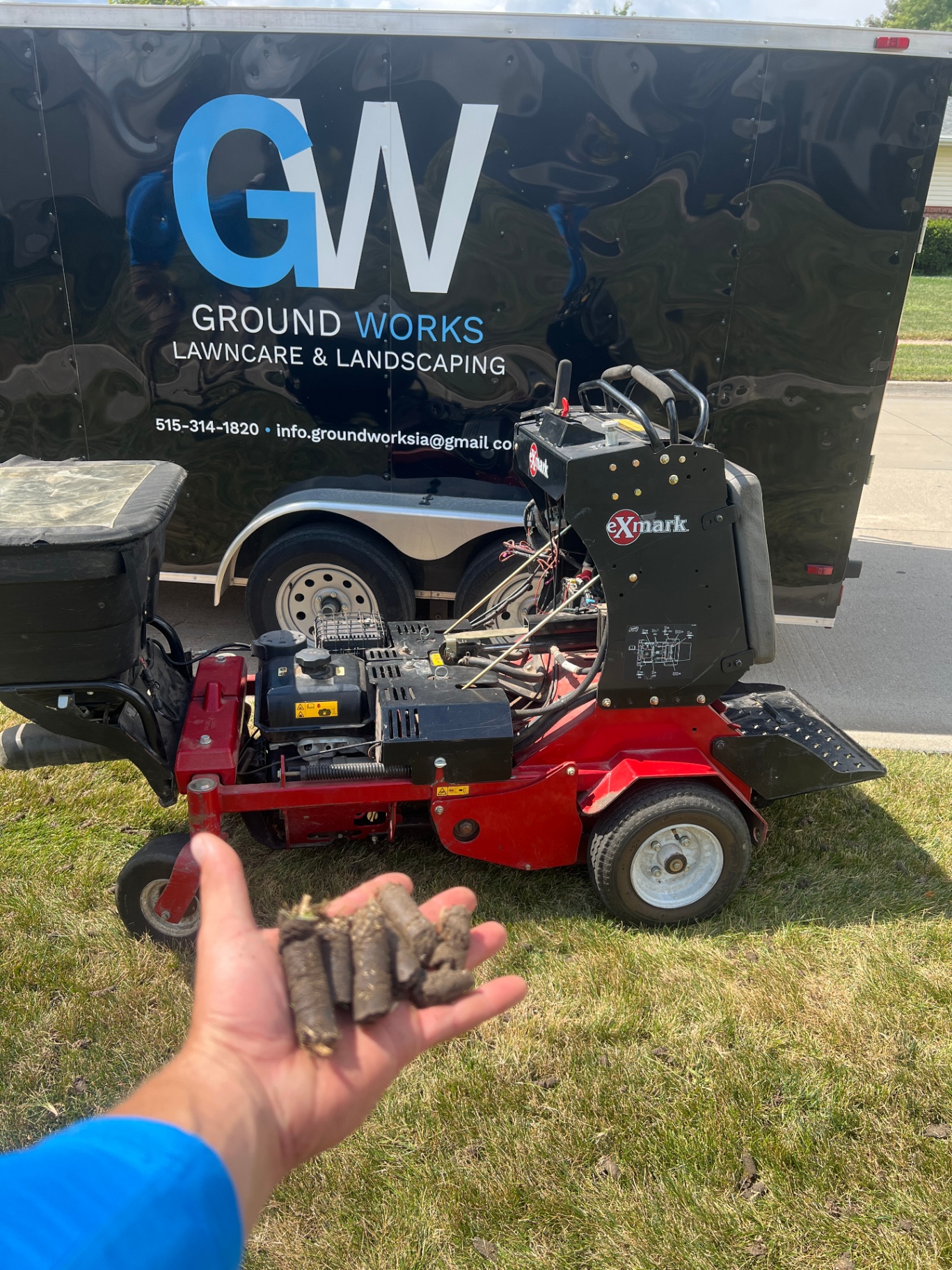 Hand holding fresh soil plugs in front of the Ground Works aerator and trailer